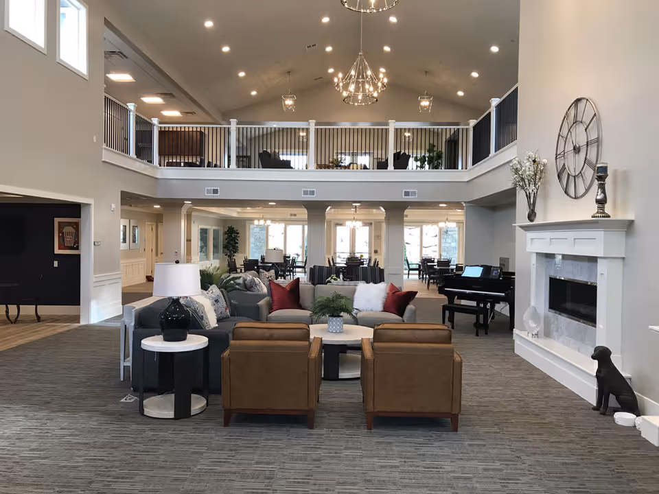 Spacious and well-lit living room area in Bellaview Assisted Living featuring a large gray sectional sofa with red and white pillows, two brown armchairs, a round coffee table, a side table with a lamp, a black piano, a white fireplace with decorative items, and a large clock on the wall. The room has high ceilings with multiple chandeliers and a second-floor balcony overlooking the space.