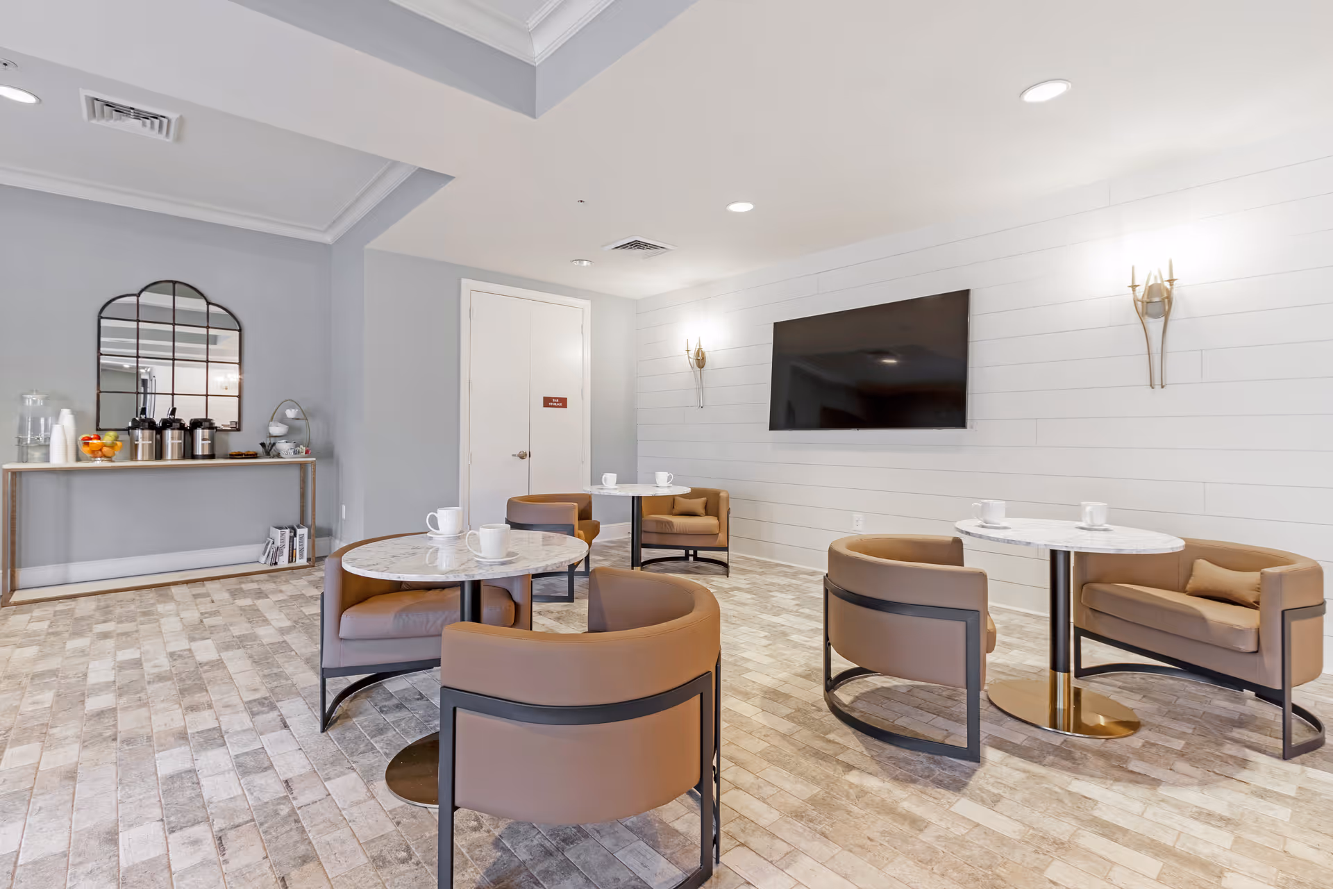 A modern lounge area with round marble-top tables and brown cushioned chairs arranged around them. The room has light-colored tiled flooring, a wall-mounted flat-screen TV, and two wall sconces. On the left side, there is a console table with coffee dispensers, cups, and a bowl of fruit beneath a large decorative mirror.