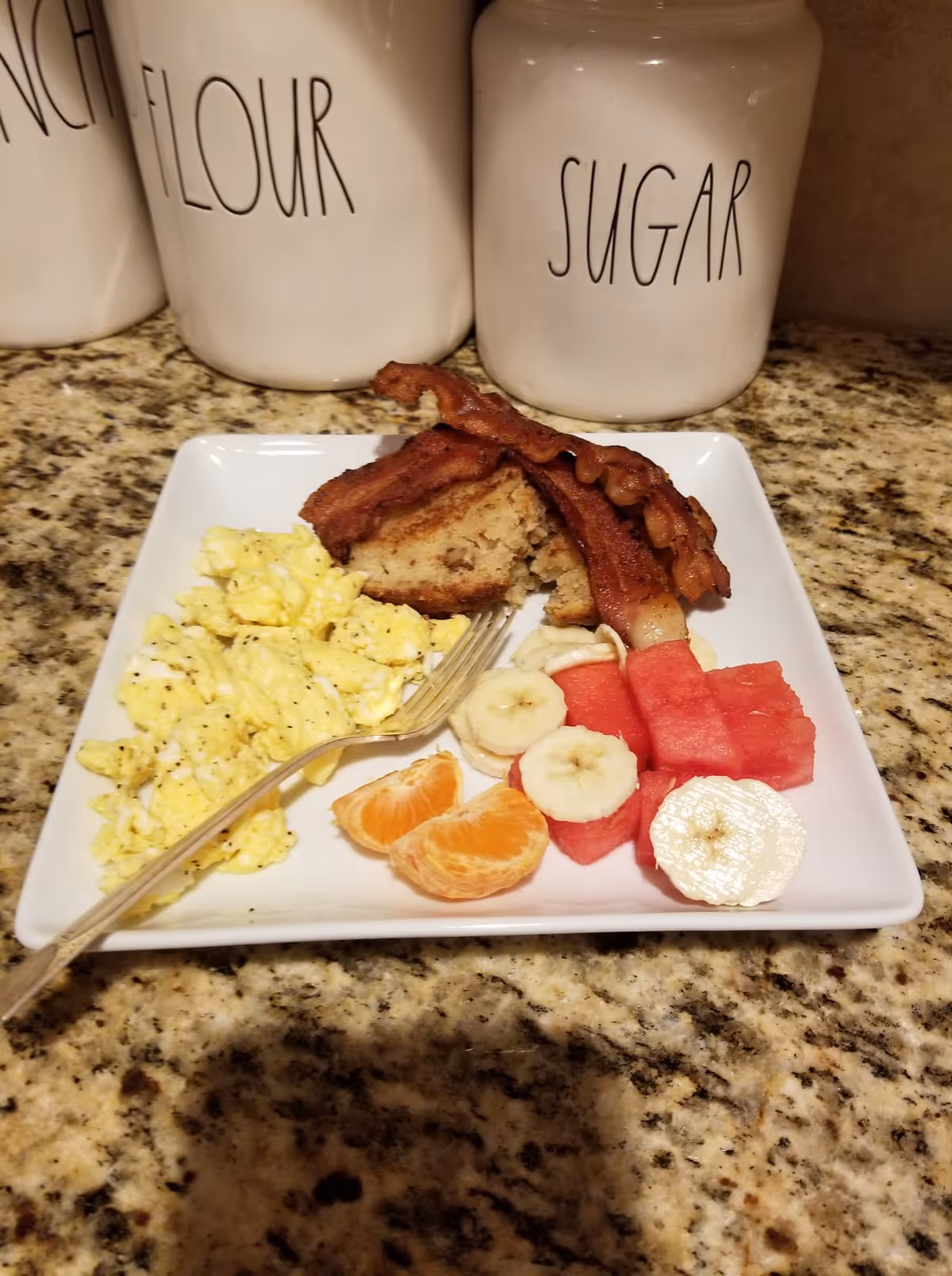 A white square plate on a granite countertop with a breakfast meal consisting of scrambled eggs, two strips of bacon, a piece of toast, slices of banana, watermelon cubes, and orange segments. Behind the plate are three white ceramic containers labeled 'FLOUR', 'SUGAR', and partially visible 'RICE'.