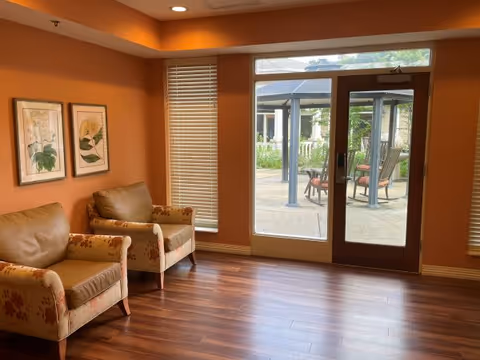 Interior sitting area with two upholstered armchairs, hardwood floors, framed botanical prints, and a glass door leading to an outdoor patio.