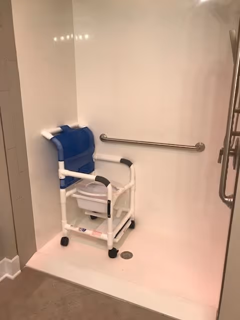 A white shower area with a blue and white shower chair equipped with wheels and a commode seat. There is a horizontal grab bar mounted on the wall for support.