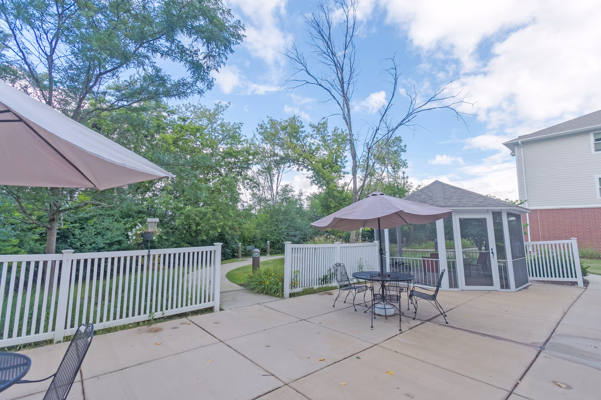Outdoor patio area with metal tables and chairs under umbrellas, a white fence, a small screened gazebo, and a paved walkway surrounded by trees and greenery under a partly cloudy sky.