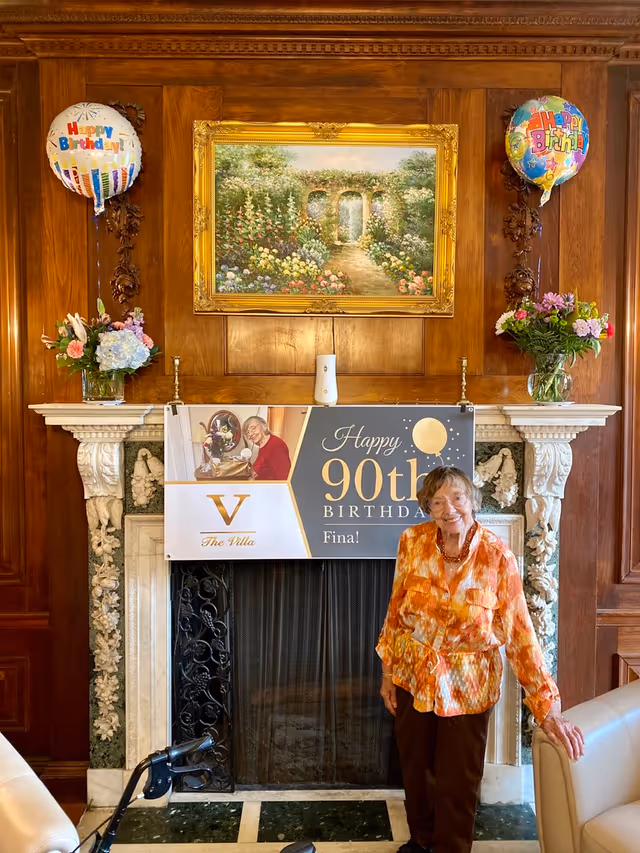 An elderly woman standing and smiling in front of a decorated fireplace with a banner that reads 'Happy 90th Birthday Fina!' The fireplace is ornate with floral carvings and a dark metal screen. Above the fireplace is a framed painting of a garden path with flowers and arches. On either side of the fireplace mantel are vases with flower arrangements and 'Happy Birthday' balloons. The room has wood-paneled walls and a beige armchair is partially visible on the right.