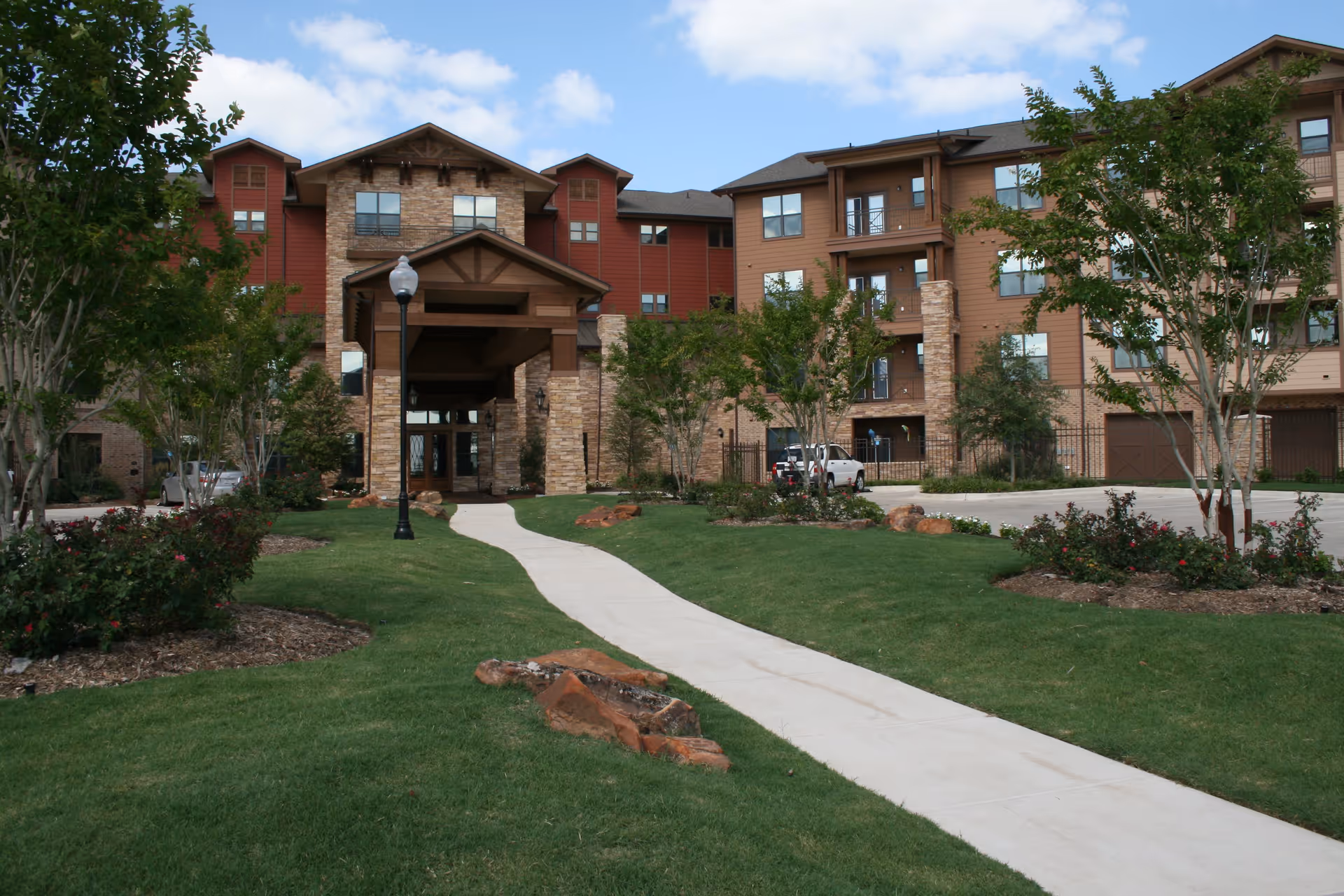Front exterior view of a senior living facility named Watercrest at Bryan, featuring a multi-story building with a covered entrance, stone and wood siding, landscaped green lawns, trees, and a paved walkway leading to the entrance under a partly cloudy sky.