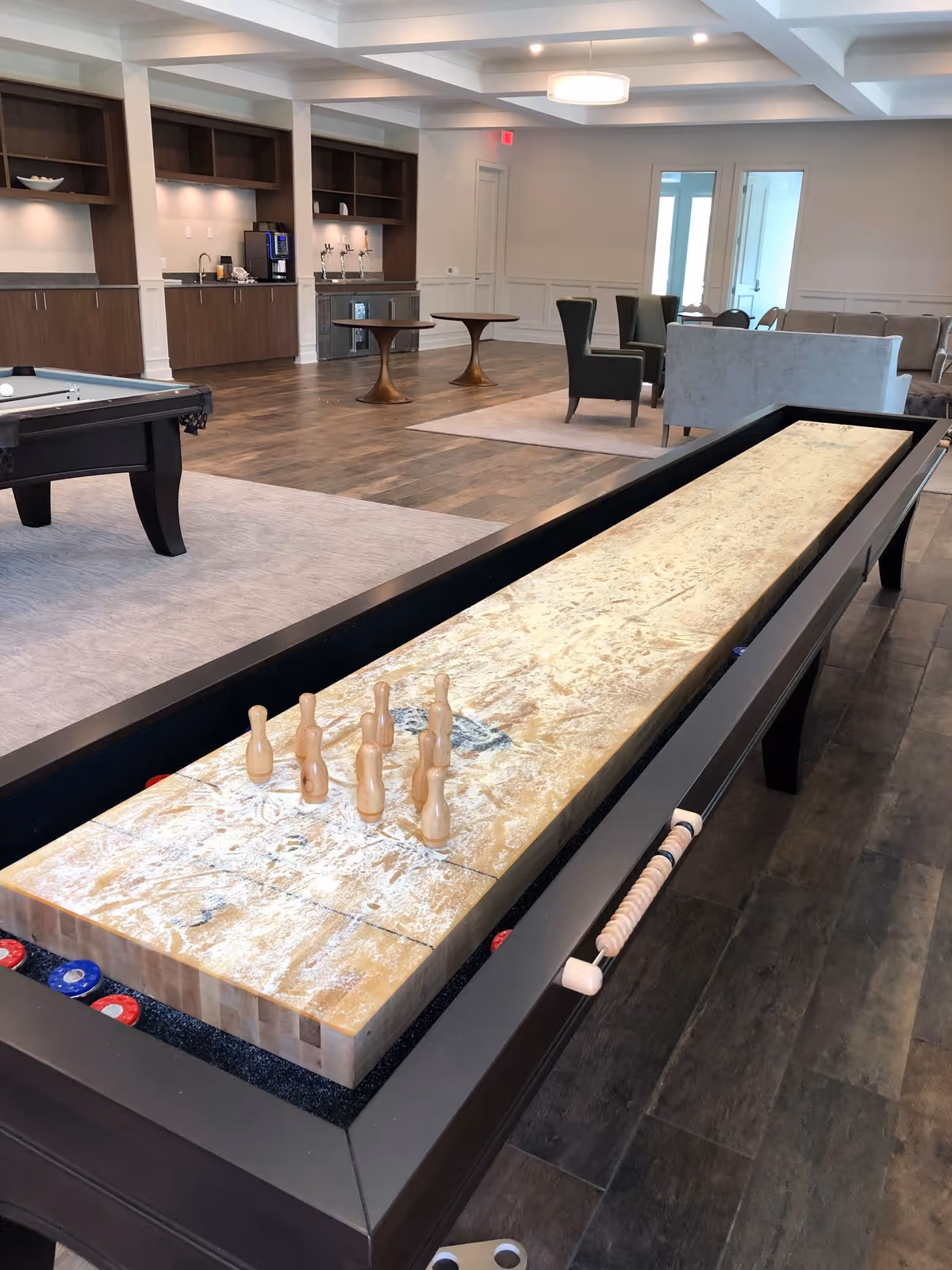 Community recreation room with a shuffleboard table in the foreground, a pool table, seating area, and a kitchenette in the background.