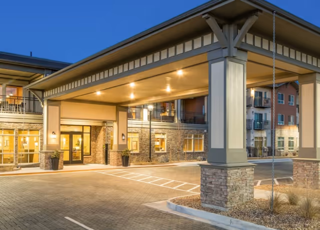 Entrance of a senior living facility at dusk with a covered drop-off area supported by large columns, illuminated by ceiling lights. The building has stone and beige siding with multiple windows and balconies visible.