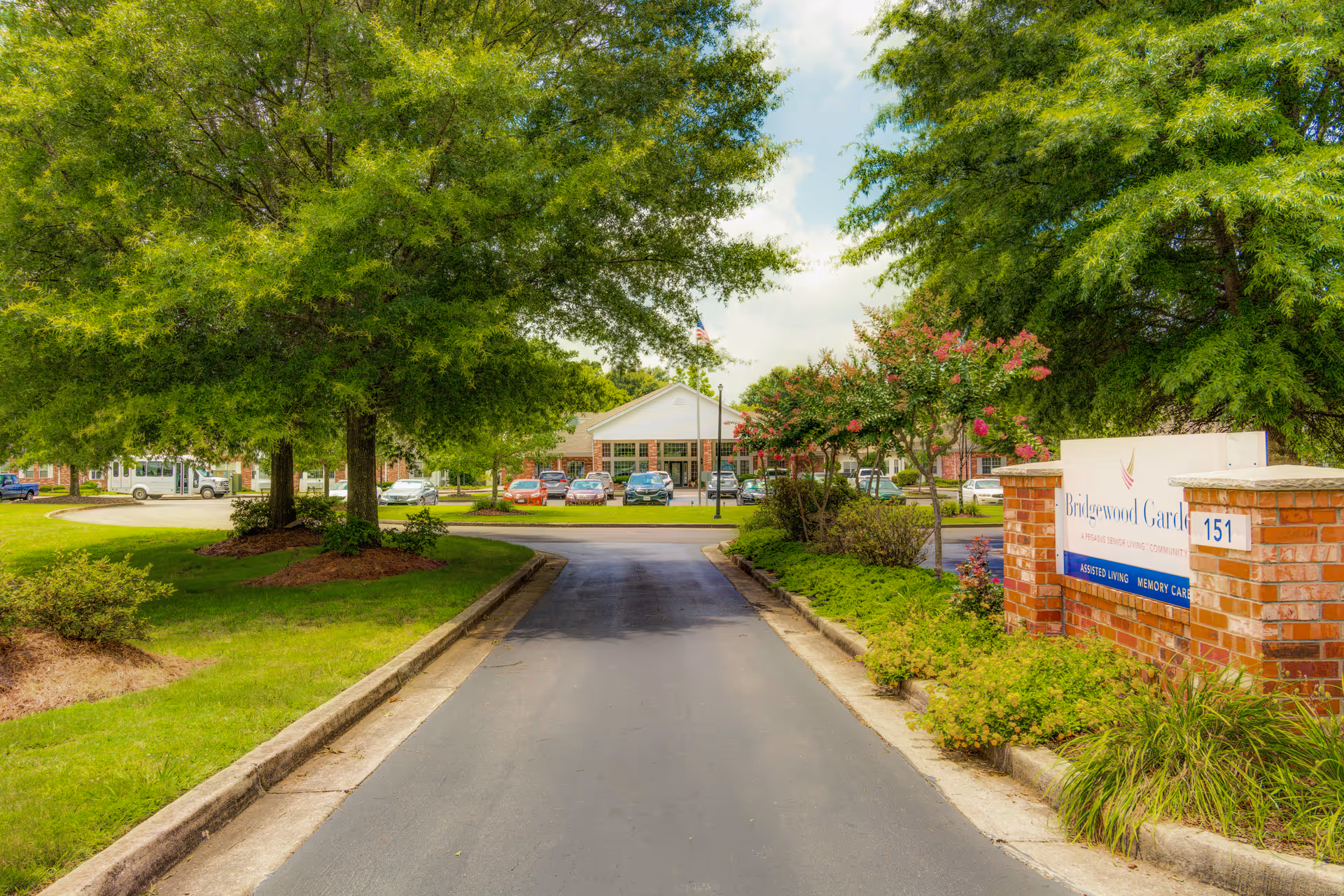 Entrance driveway leading to the main building of Bridgewood Gardens, a senior living community, with green trees and landscaped bushes on both sides. A sign with the facility name and address number 151 is visible on the right side near the entrance.