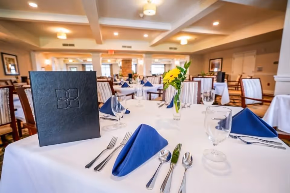 A dining room with tables covered in white tablecloths, set with blue folded napkins, silverware, water glasses, and a small vase with a yellow flower. The room has wooden chairs with striped cushions and soft lighting from ceiling fixtures.