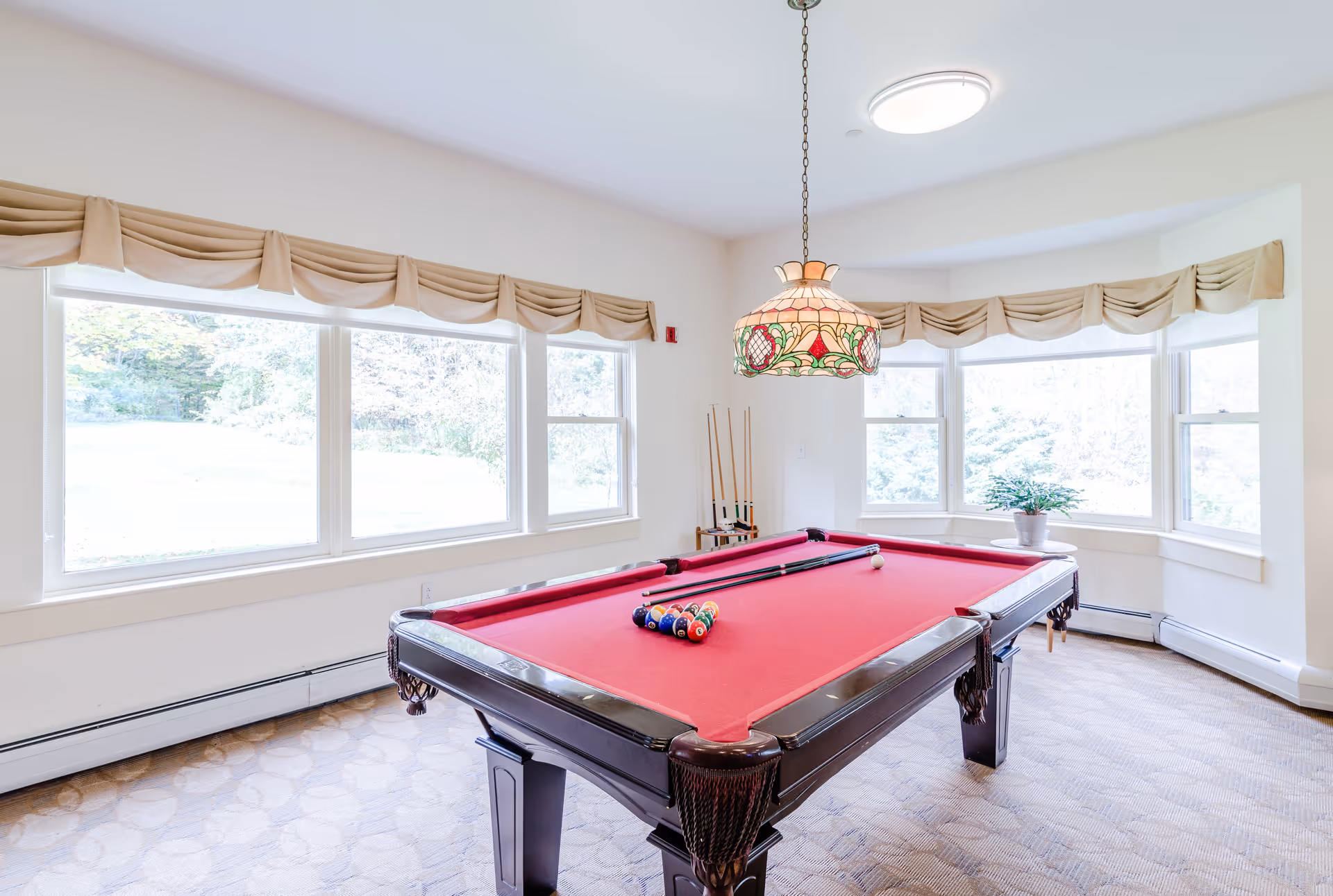 Bright game room with a red-felt pool table centered beneath a stained-glass pendant lamp and large bay windows.
