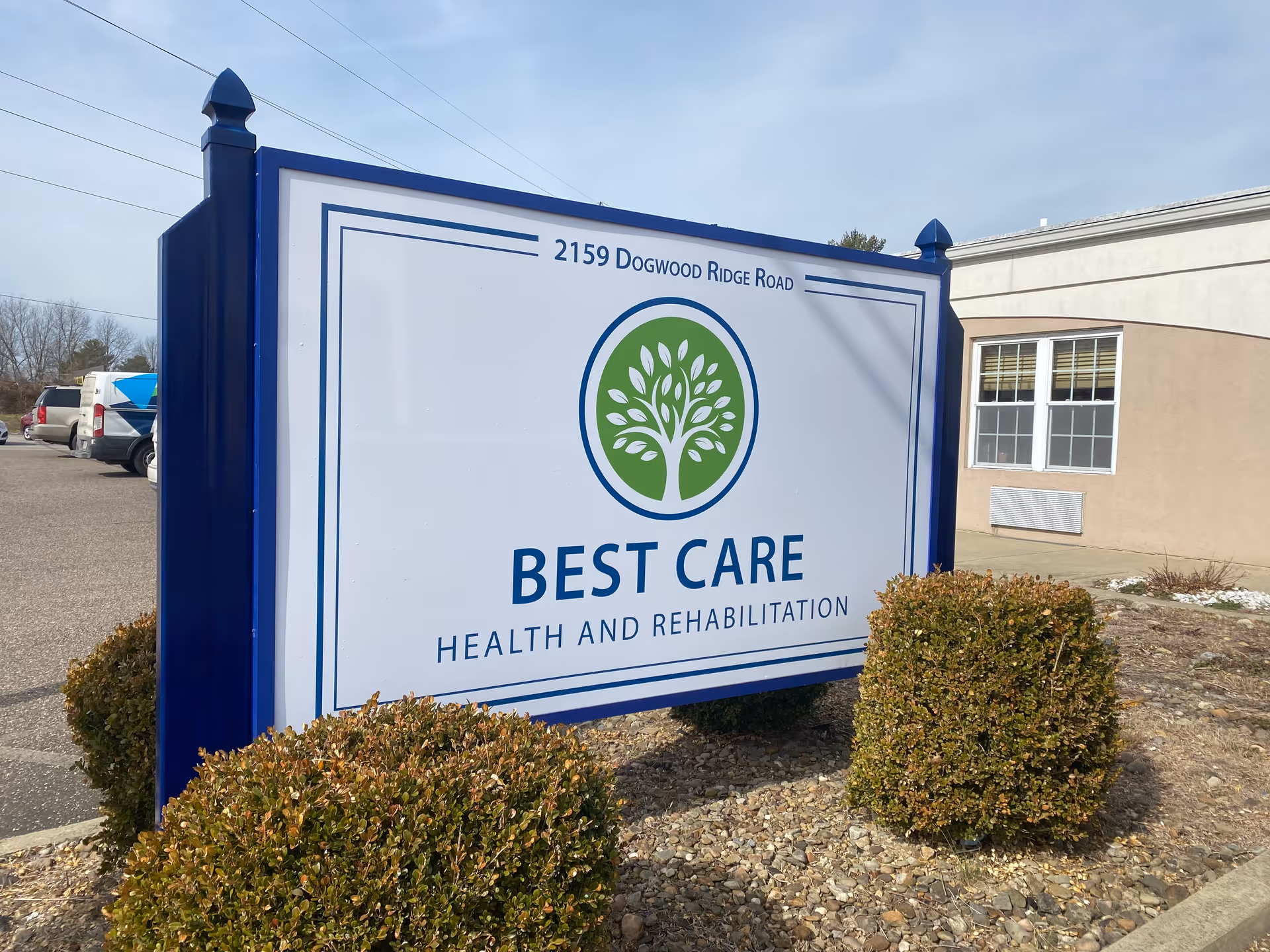 Outdoor image of a large sign for Best Care Health and Rehabilitation located at 2159 Dogwood Ridge Road, surrounded by bushes and a parking lot with vehicles in the background.
