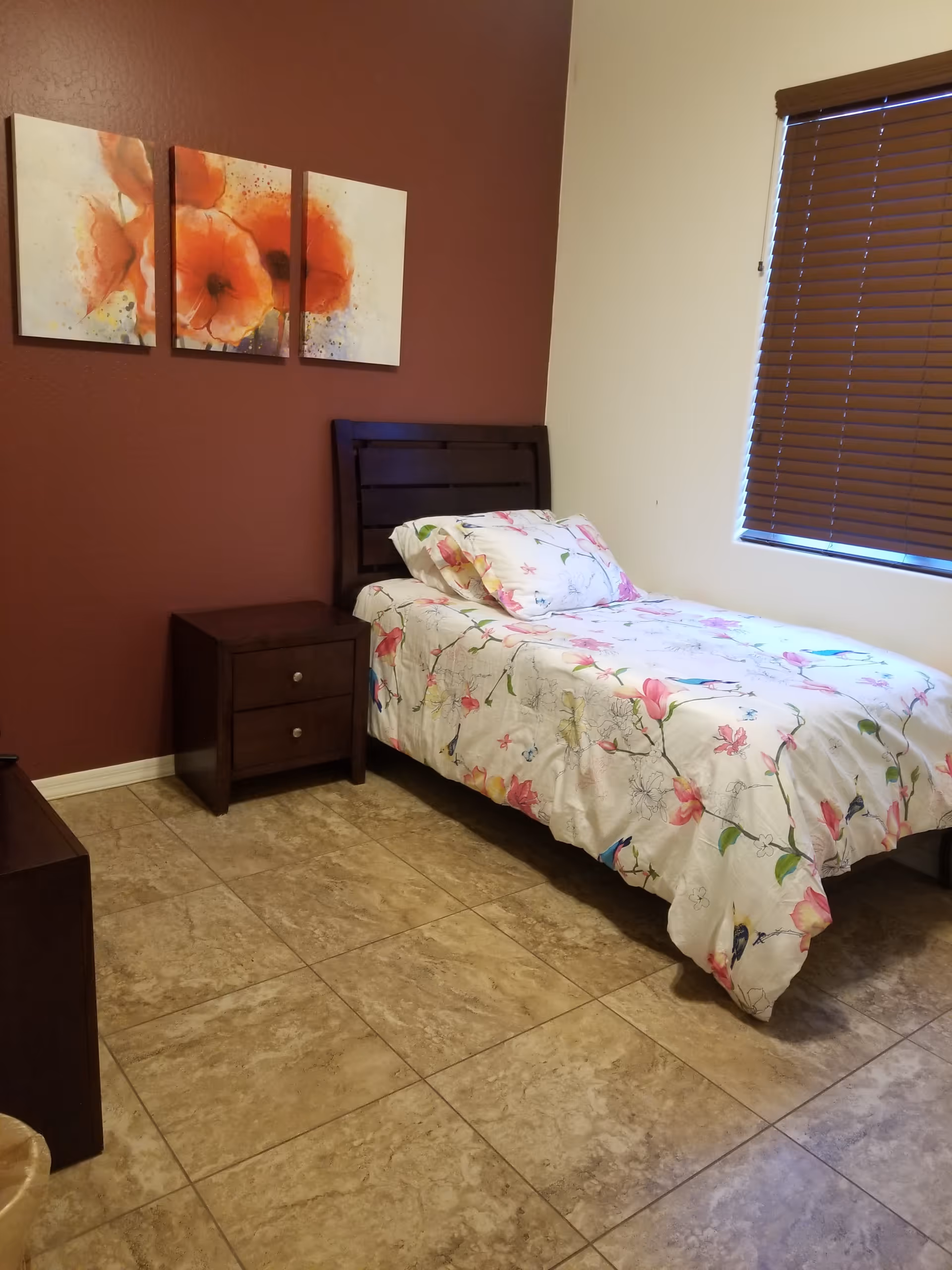 Small bedroom with a single bed in floral bedding, a wooden headboard and nightstand, floral wall art, and a window with blinds.
