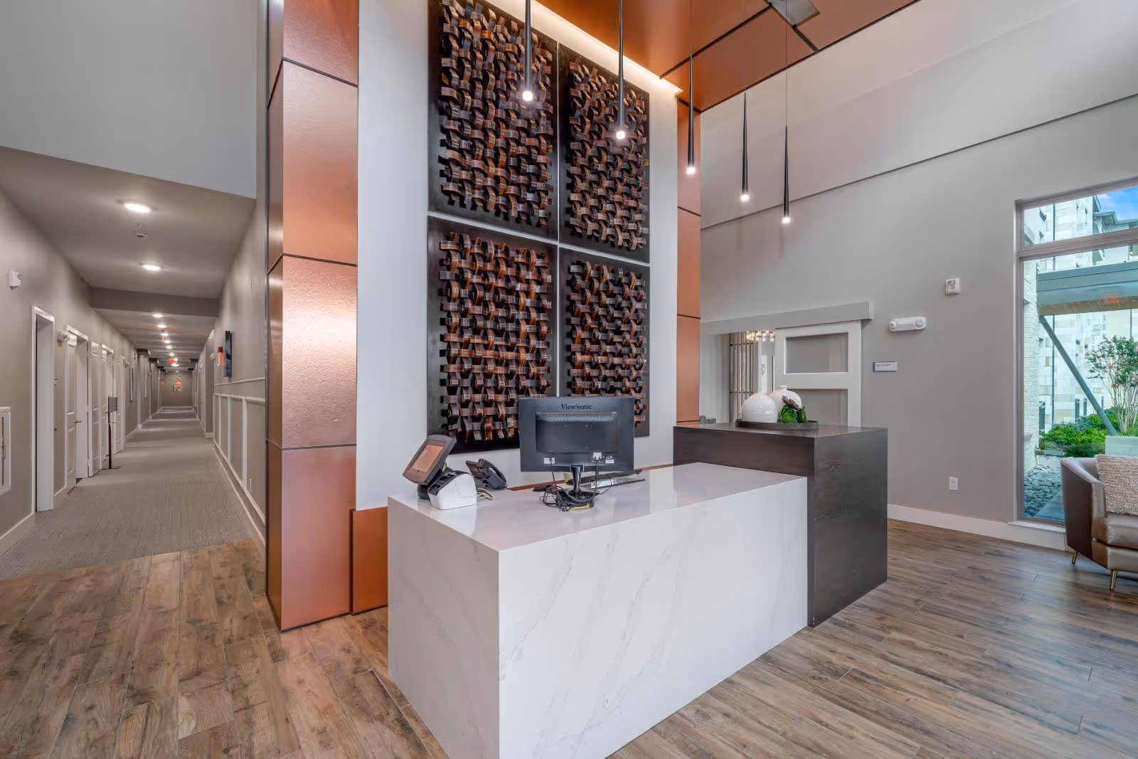 Modern reception area with a white marble front desk and a dark wood side counter, featuring a computer monitor and a phone. Behind the desk is a tall decorative wall art with four panels of woven dark wood. Pendant lights hang from the ceiling above the desk. To the left, a long hallway with multiple doors and ceiling lights extends into the distance. To the right, a large window shows an outdoor view with greenery and part of a building. The floor is a mix of wood and carpet.