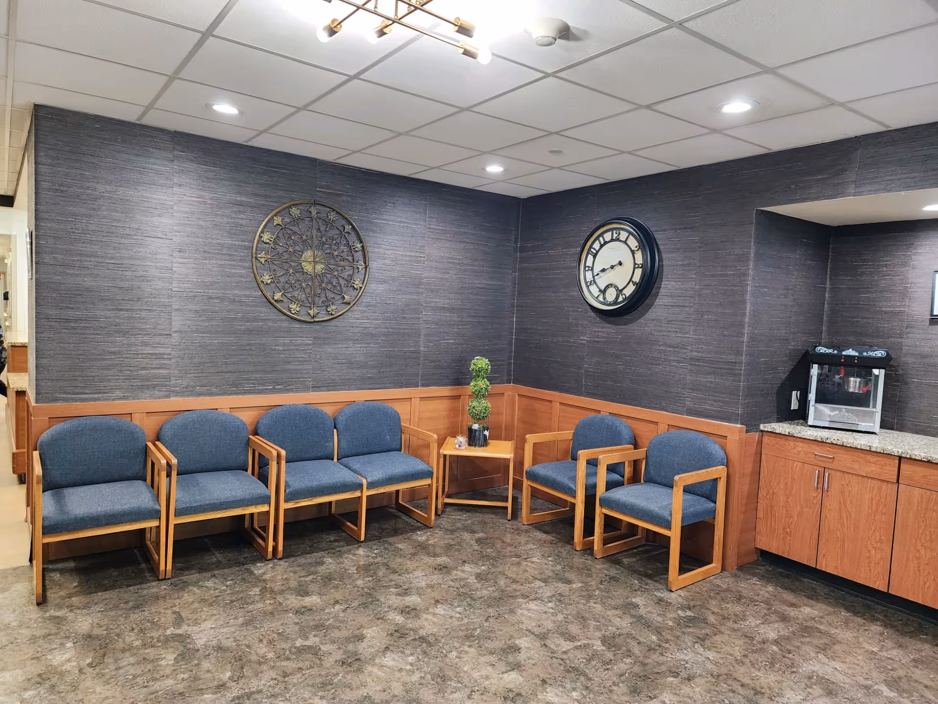 A waiting area with five blue cushioned wooden chairs arranged along two walls, a small wooden corner table with a decorative plant, a large round wall clock, and a decorative metal wall art piece. There is a popcorn machine on a granite countertop with wooden cabinets below. The floor has a stone-like pattern and the ceiling has recessed lighting.