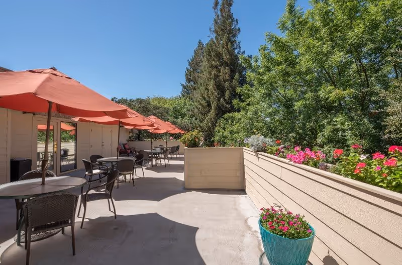 Outdoor patio area with several round tables and chairs under red umbrellas. The patio is surrounded by a beige wooden fence with flower boxes containing pink and red flowers. There are tall green trees and a clear blue sky in the background.