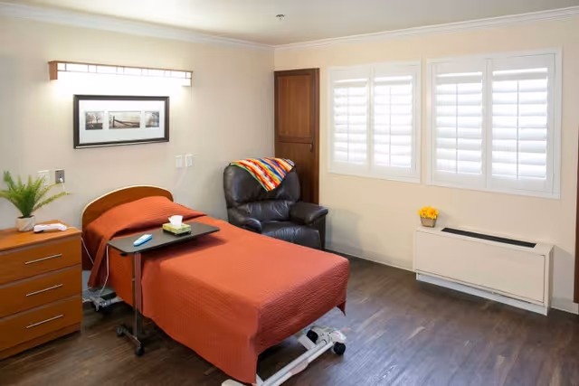 A small bedroom with a single hospital-style bed covered with a rust-colored blanket, a black leather recliner with a colorful blanket draped over the back, a wooden dresser with a potted plant and a book, a wall-mounted light fixture above a framed picture, and windows with white shutters letting in natural light.