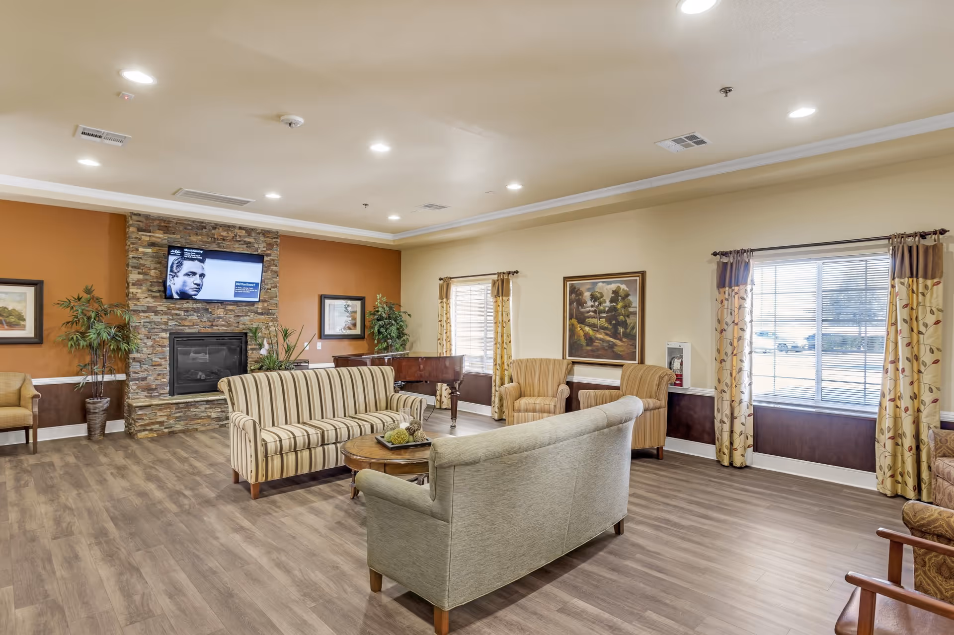 A spacious living room with wood flooring, beige walls, and recessed lighting. The room features a stone fireplace with a mounted flat-screen TV above it, two striped sofas facing each other around a round wooden coffee table, two armchairs near a window with patterned curtains, and a grand piano in the corner. There are framed paintings on the walls and potted plants adding greenery to the space.