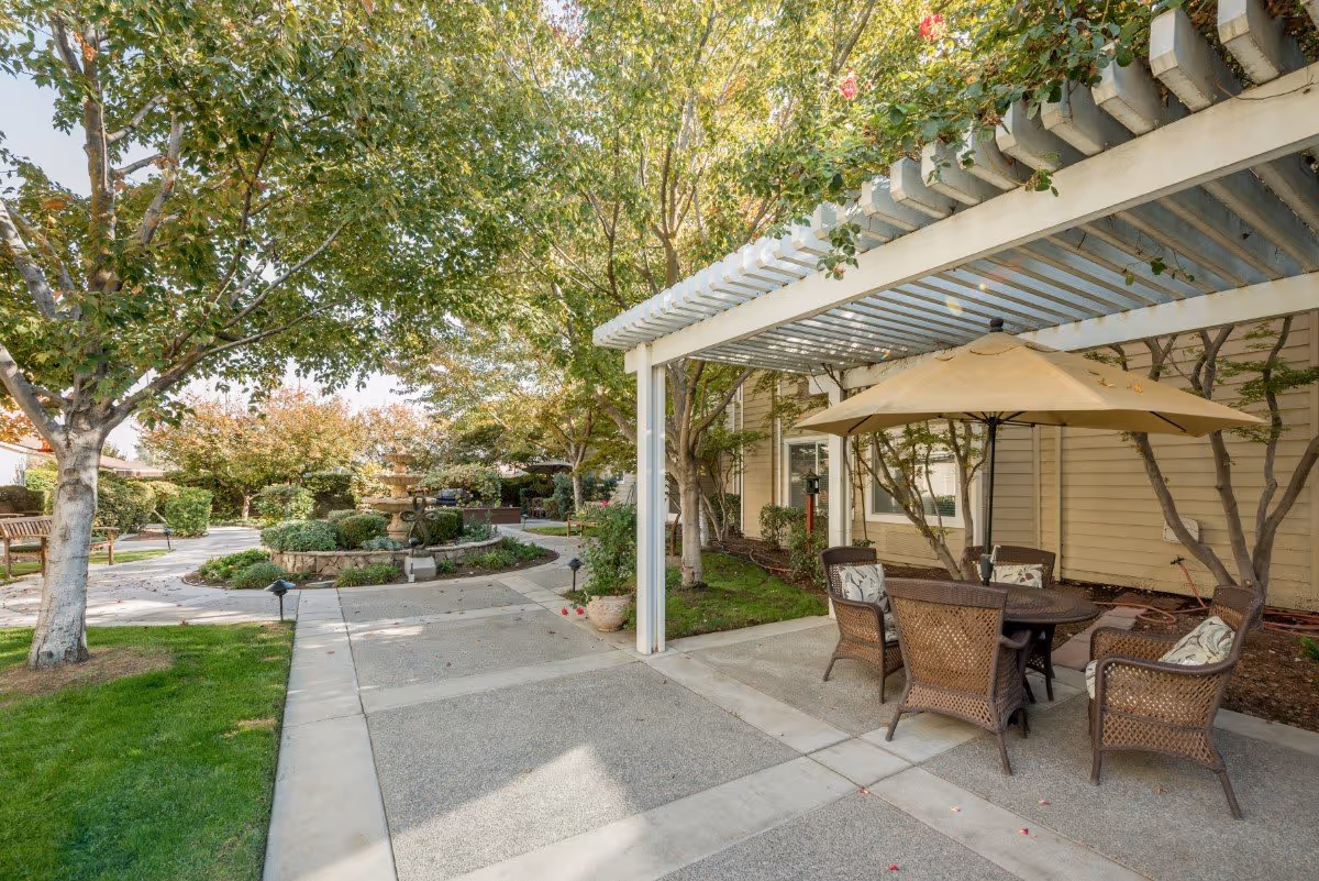 Outdoor patio area at The Grove Senior Living with a round table and five wicker chairs under a large beige umbrella. The patio is shaded by a white pergola and surrounded by trees and greenery. A stone fountain and benches are visible in the background along a paved walkway.