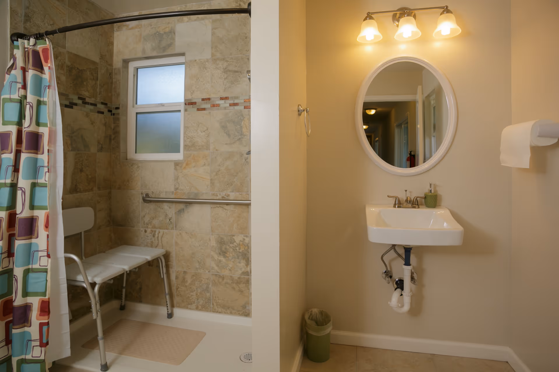 A bathroom with a tiled walk-in shower featuring a white shower chair and a colorful shower curtain. Next to the shower is a wall-mounted sink with a round mirror above it, three light fixtures, a soap dispenser, a towel ring, a toilet paper holder with a roll of toilet paper, and a small trash can on the floor.