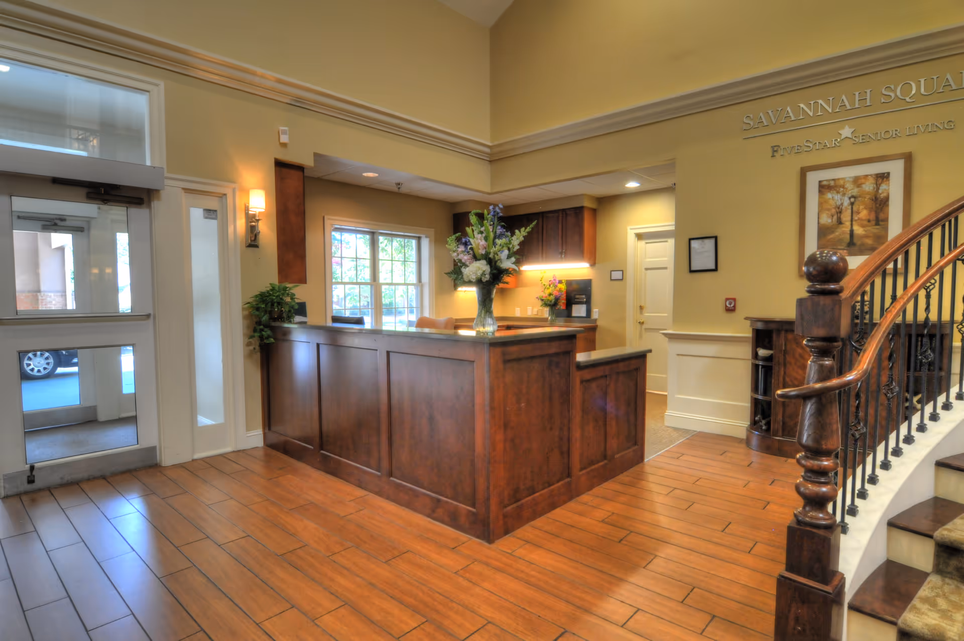 Reception desk and entry lobby of Savannah Square senior living with a wooden counter, floral arrangement, and a curved staircase.