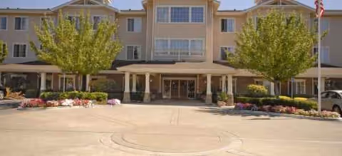 Senior living facility with a beige building, entrance, and landscaped trees in front.