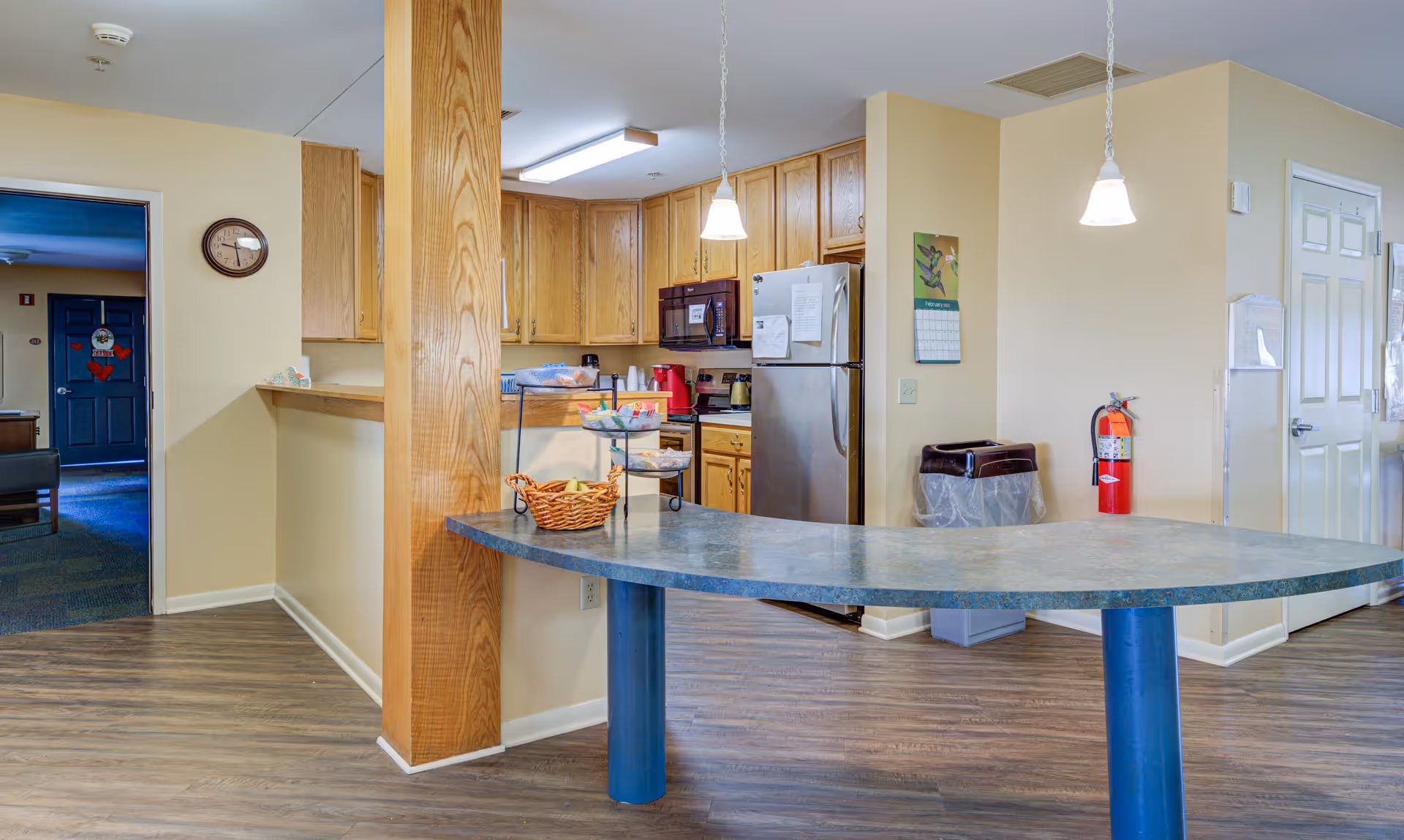 Interior view of a senior living community kitchen area with wooden cabinets, a stainless steel refrigerator, microwave, and a curved countertop with a basket of fruit and tiered trays. The floor is wood-style laminate, and there are two hanging pendant lights above the counter. A fire extinguisher and trash bins are visible against the wall, with a doorway leading to another room in the background.