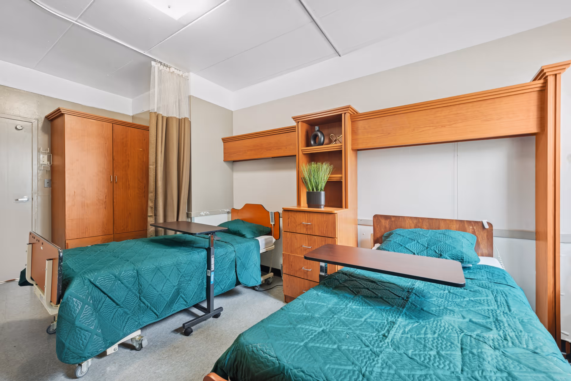 A shared patient room with two hospital-style beds with green bedspreads, wooden storage units, and overbed tables.