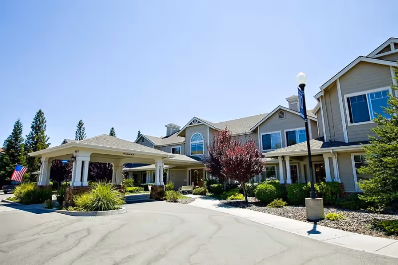 Exterior view of a senior living facility building with a covered entrance driveway, landscaped greenery, trees, and a clear blue sky.