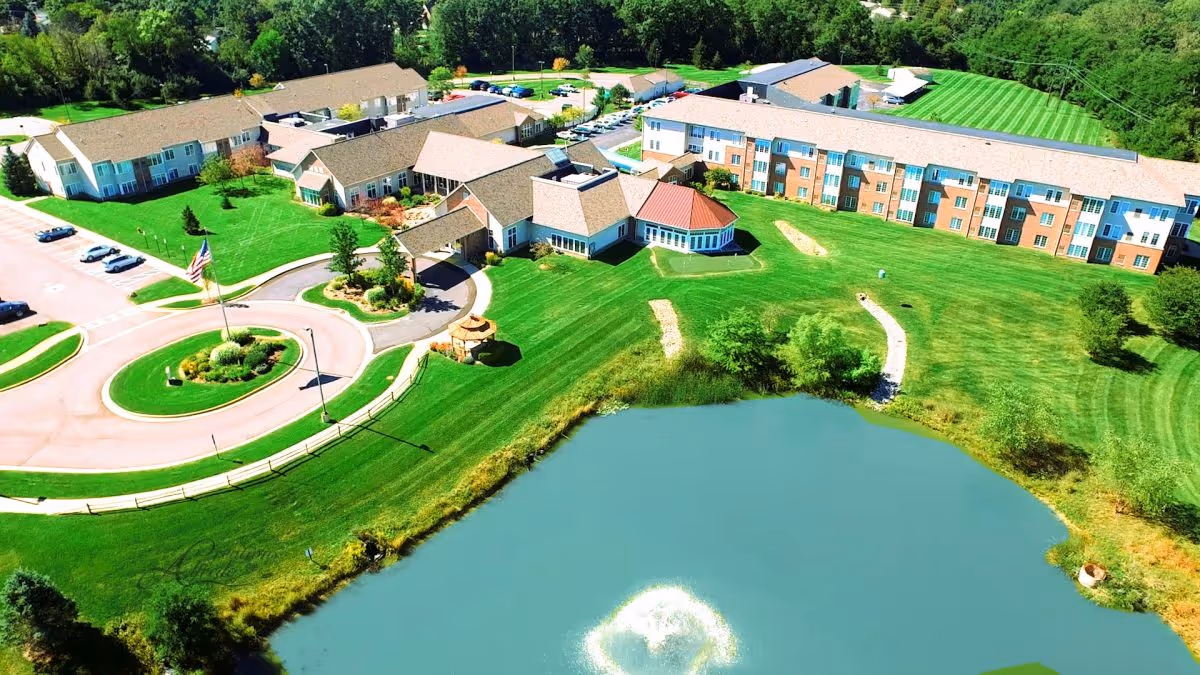 Aerial view of Brecon Village senior living facility showing multiple connected buildings with beige and brick exteriors, a circular driveway with an American flag in the center, well-maintained green lawns, a small pond with a water fountain, and surrounding trees.