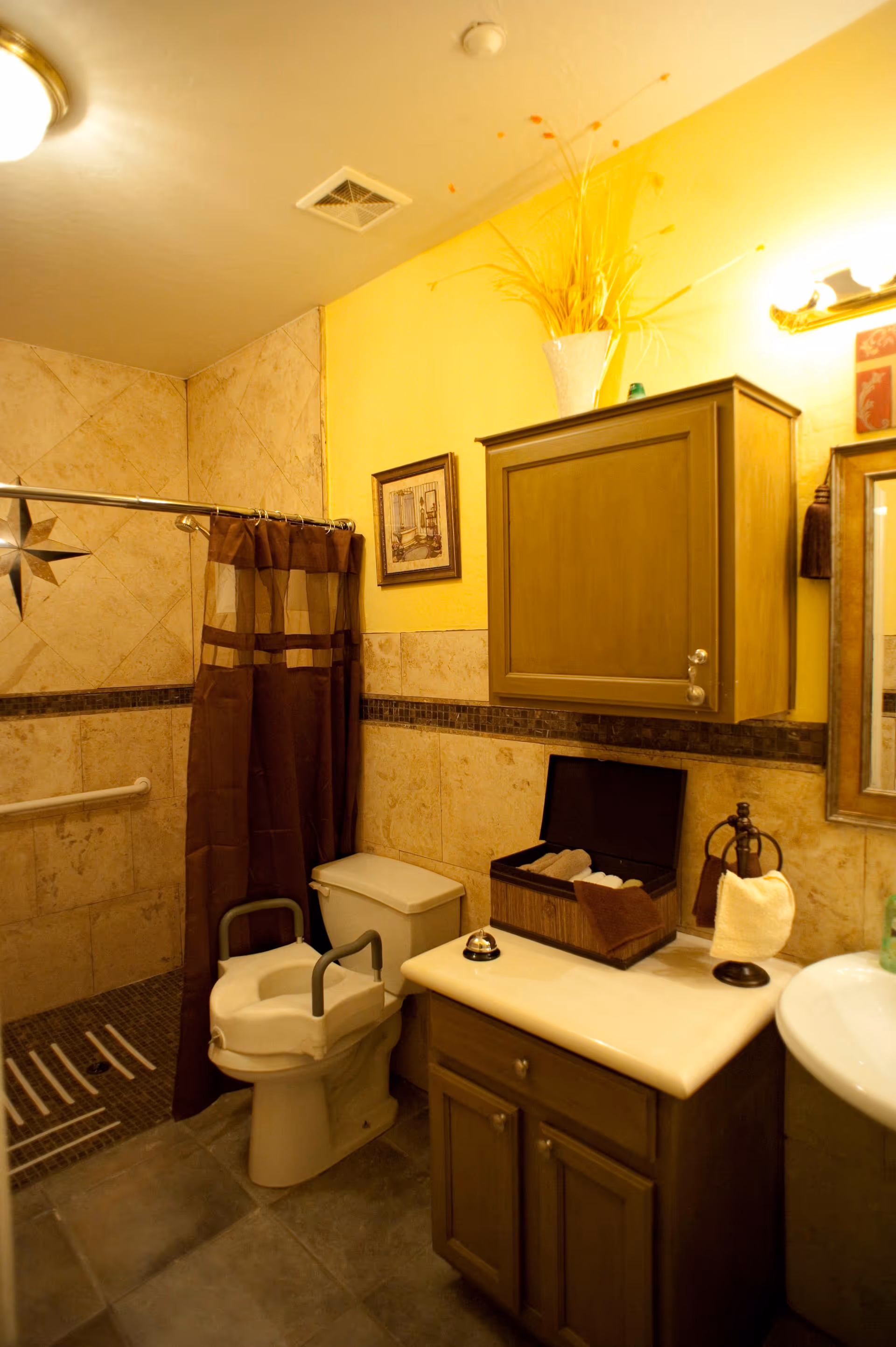 A bathroom with beige tiled walls and floor, featuring a toilet with a raised seat and support handles, a shower area with a brown curtain, a wooden cabinet mounted on the wall, a countertop with a box of rolled towels, a small bell, and a towel holder with a white towel. The walls are painted yellow above the tiles, and there is a framed picture and a vase with decorative grass on top of the cabinet.