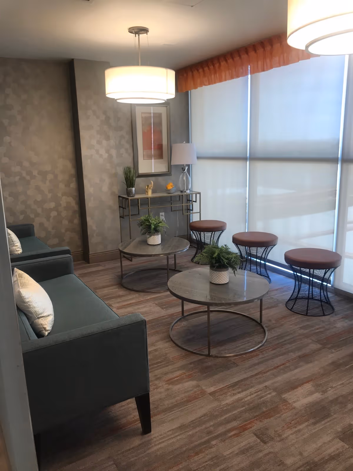 A modern living room area with two green sofas, two round coffee tables with potted plants, three round stools with brown cushions, a console table with decorative items and a lamp, and large windows covered with white roller blinds.