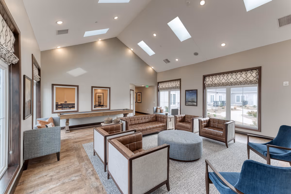 A spacious and well-lit senior living facility common area with high vaulted ceilings and skylights. The room features a seating arrangement with brown leather sofas and chairs, blue upholstered chairs, a large round ottoman in the center, and a long wooden shuffleboard table against the far wall. Large windows with patterned valances allow natural light to enter, and the flooring is a combination of wood and carpet.