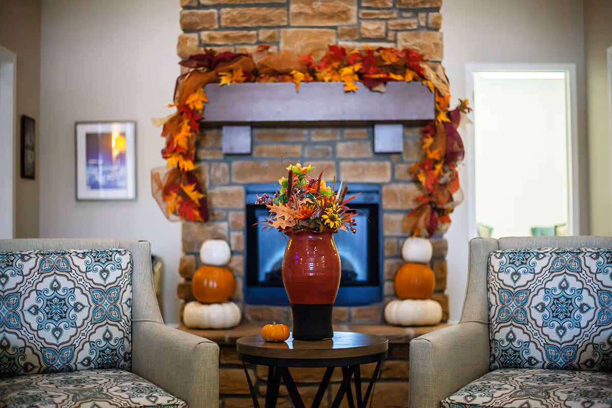 Cozy living room area with two patterned armchairs facing a small round table that holds a red vase filled with autumn-themed flowers and foliage. Behind the table is a stone fireplace decorated with a garland of fall leaves and pumpkins stacked on either side.