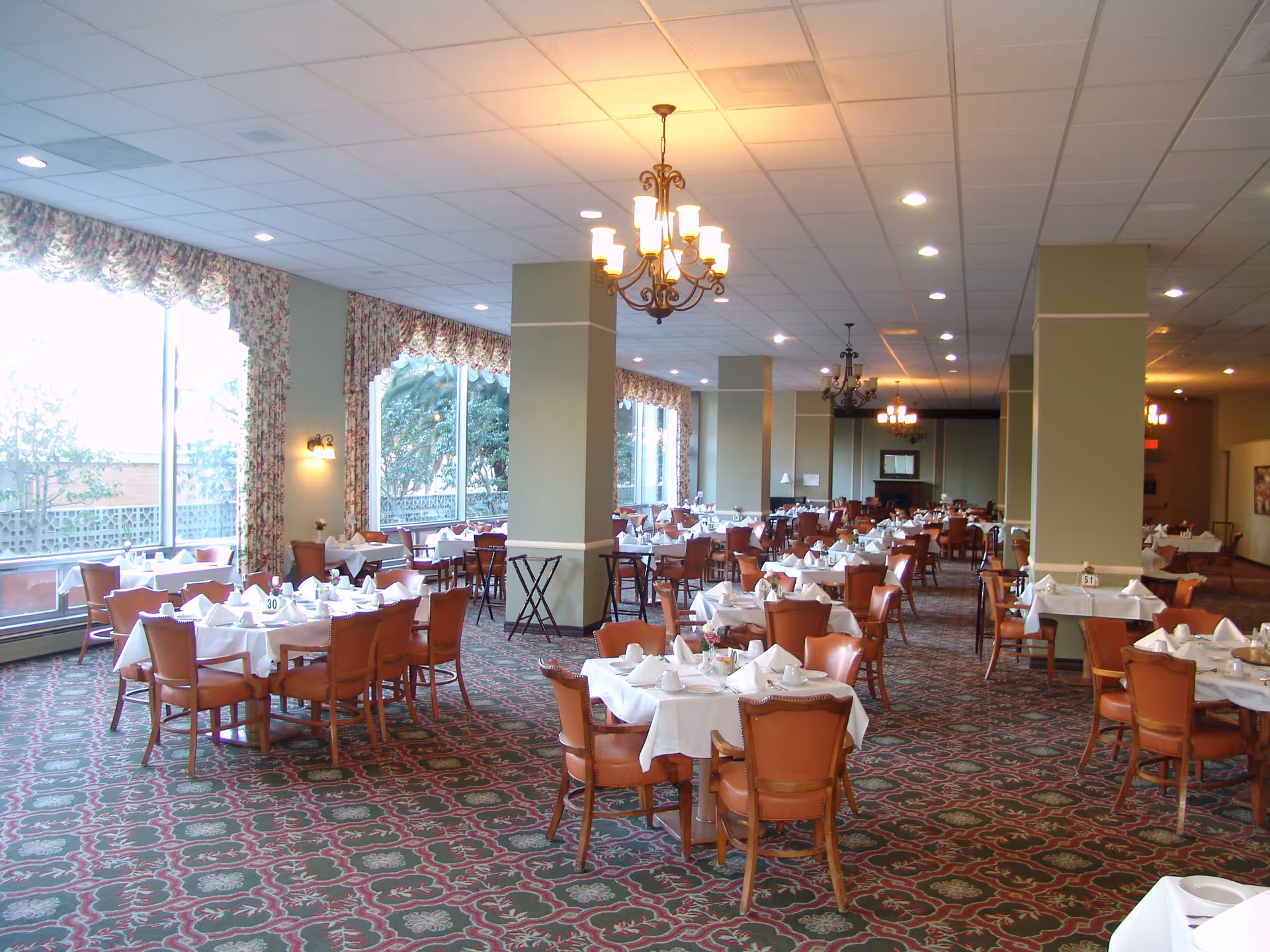 A spacious dining room with multiple tables covered in white tablecloths and set with napkins, cups, and plates. The room has large windows with floral curtains, green walls with columns, patterned carpet, and several chandeliers hanging from the ceiling.