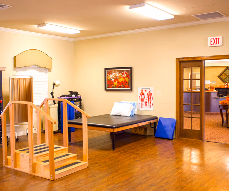 A rehabilitation therapy room with wooden stairs for physical therapy, a black padded therapy table with pillows, a medical privacy screen, anatomical chart on the wall, and a framed floral painting. The room has wooden flooring and beige walls, with a doorway leading to another room.