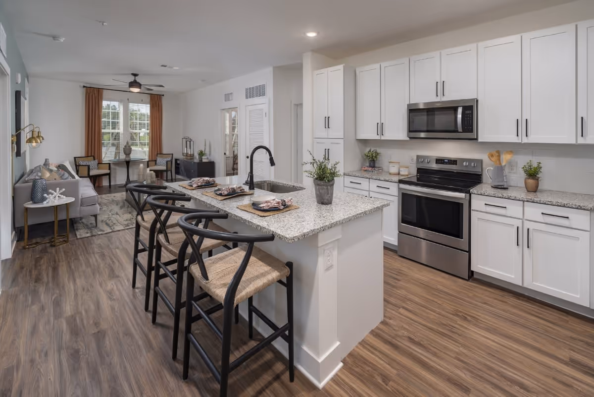 Modern open-concept kitchen and living area with white cabinets, stainless steel appliances, a granite countertop island with four black bar stools, and a small dining table with two chairs near a window with brown curtains.