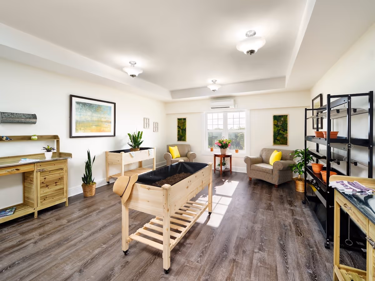 A bright, spacious room with wooden flooring featuring two beige armchairs with yellow cushions near a window. The room contains wooden gardening tables with soil, a black metal shelving unit with empty terracotta pots, and various green plants. The walls are white with framed artwork and two vertical green plant displays. The ceiling has recessed lighting fixtures.