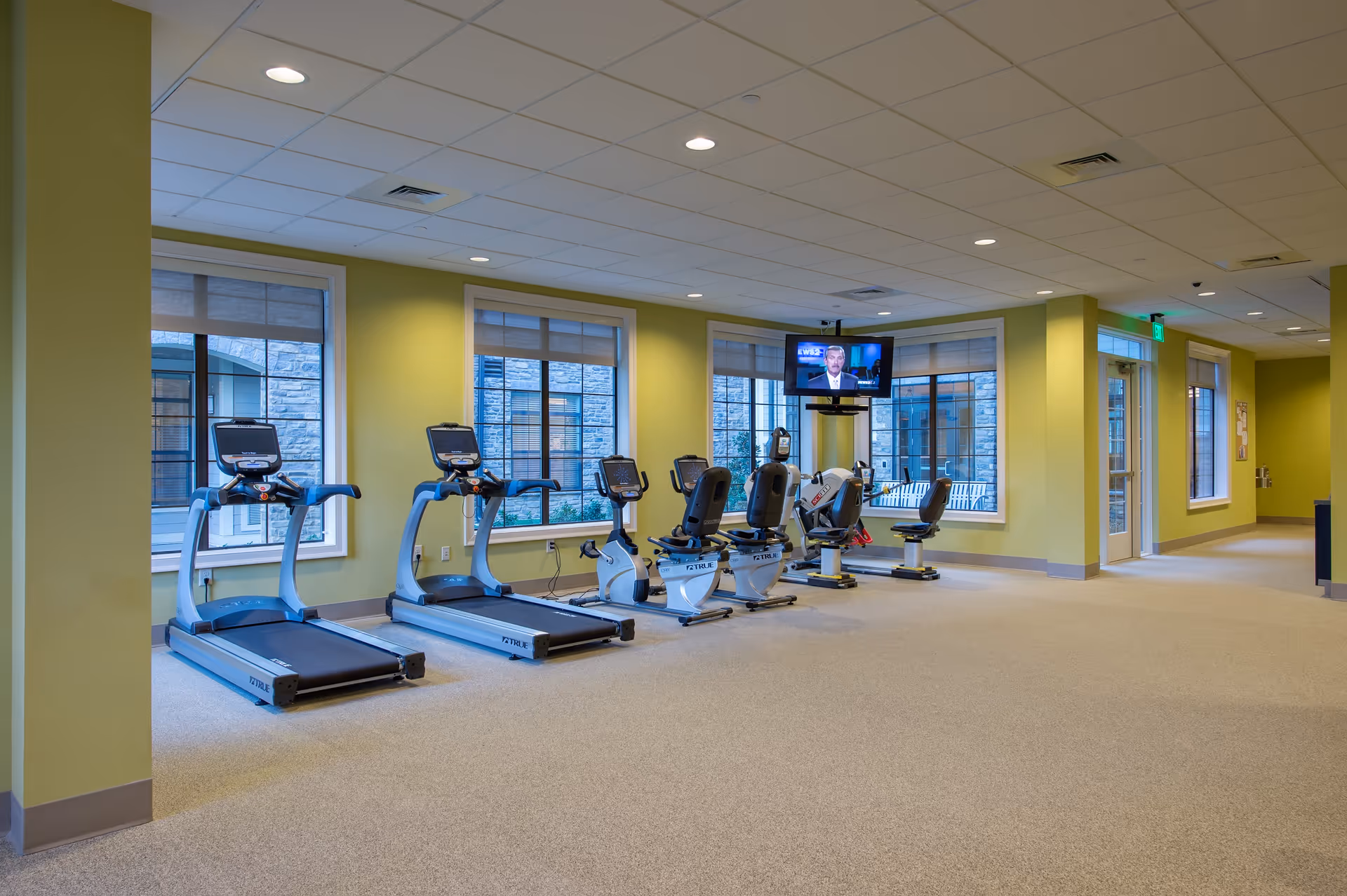 A fitness room in a senior living facility with two treadmills, three stationary bikes, and a television mounted on the wall. The room has large windows with views of an outdoor area, light green walls, and a beige carpeted floor.