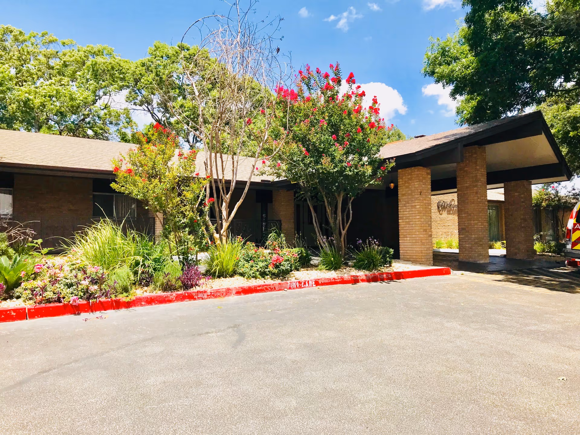 Exterior view of a single-story brick building with a covered entrance supported by brick columns. There are flowering trees and shrubs in front of the building, a clear blue sky with some clouds, and a paved driveway with a red curb marked 'FIRE LANE'.
