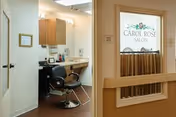 Interior view of a salon area in a senior living facility. The image shows a salon chair in front of a counter with a mirror, hair care products, and a sink. To the right, there is a door with a frosted glass panel displaying the text 'Carol Rose Salon' with a floral design above it.