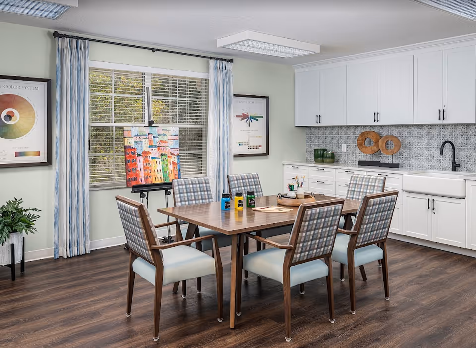 A bright and clean art activity room with a wooden table surrounded by six cushioned chairs with plaid backs. On the table are paint bottles, brushes, and art supplies. Behind the table is a large window with blue and white curtains, an easel holding a colorful painting, and two framed color system charts on the walls. The room features white cabinetry, a farmhouse sink, and a patterned tile backsplash.