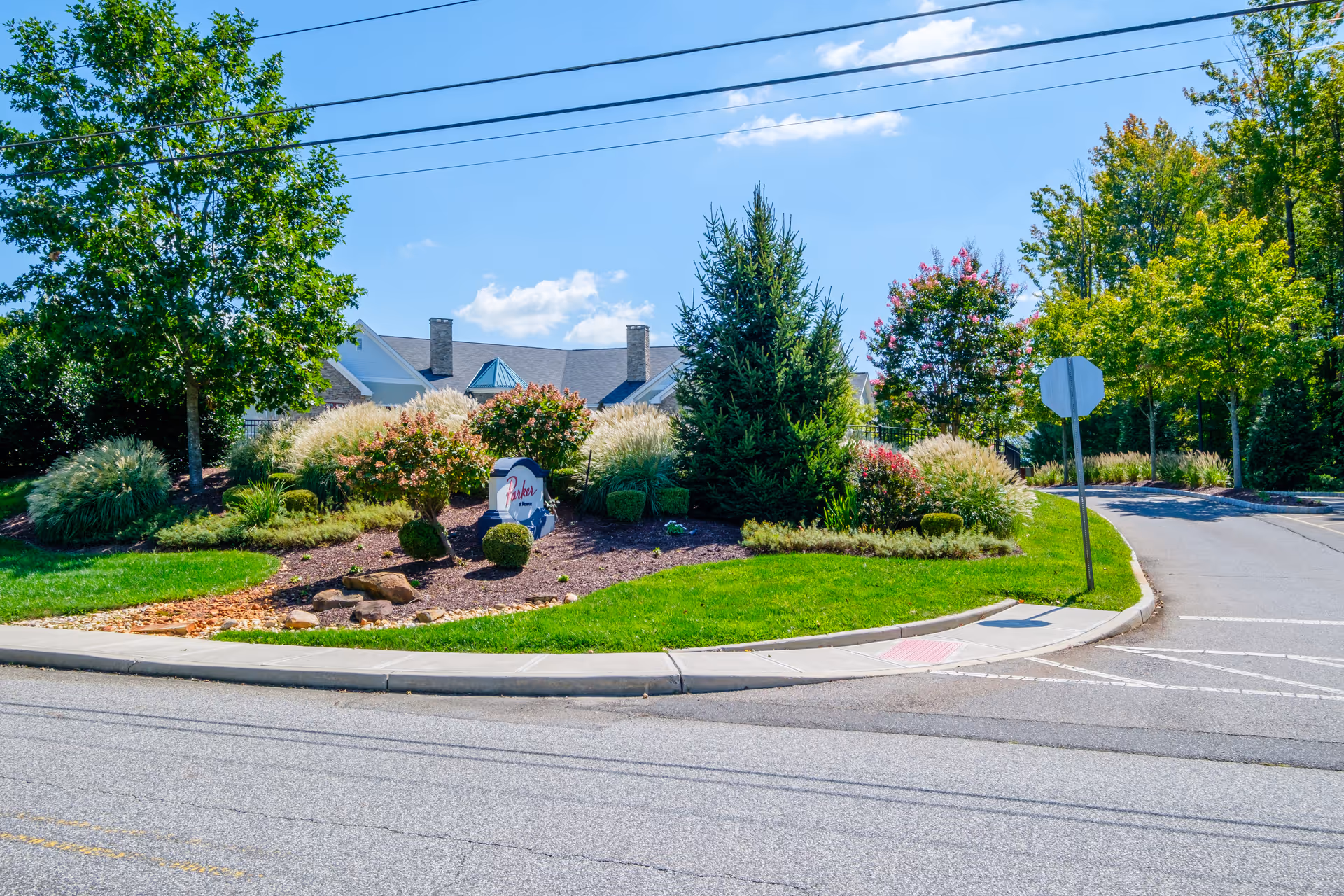 A landscaped entrance area with green grass, bushes, trees, and a sign that reads 'Parker at Monroe' under a clear blue sky with some clouds. A road curves around the landscaped area with a stop sign visible.