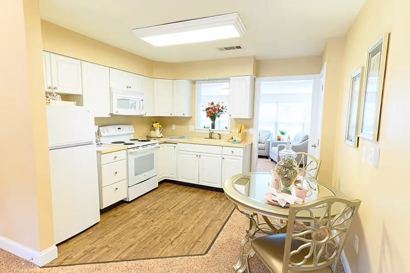 A bright kitchen area with white cabinets, a white refrigerator, stove, and microwave. There is a small round glass dining table with two chairs on the right side. The kitchen floor is wood, transitioning to carpet in the dining area. A window above the sink shows a vase with flowers. In the background, a living room with a sofa and large windows is visible.