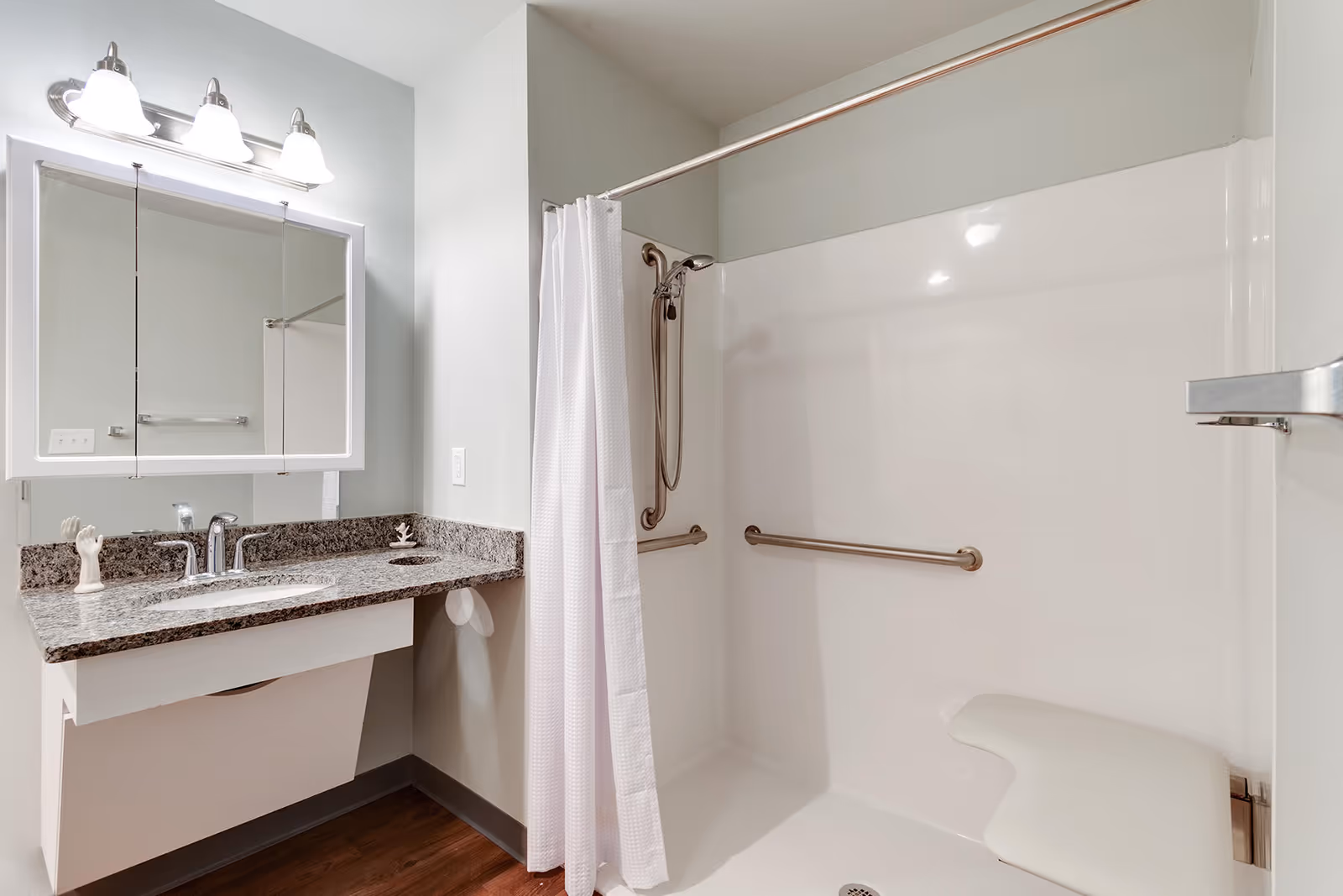 A clean and modern bathroom with a granite countertop sink, a large mirror with three overhead lights, and a walk-in shower equipped with grab bars, a handheld showerhead, and a built-in seat. The shower has a white curtain and the floor is wood-style.
