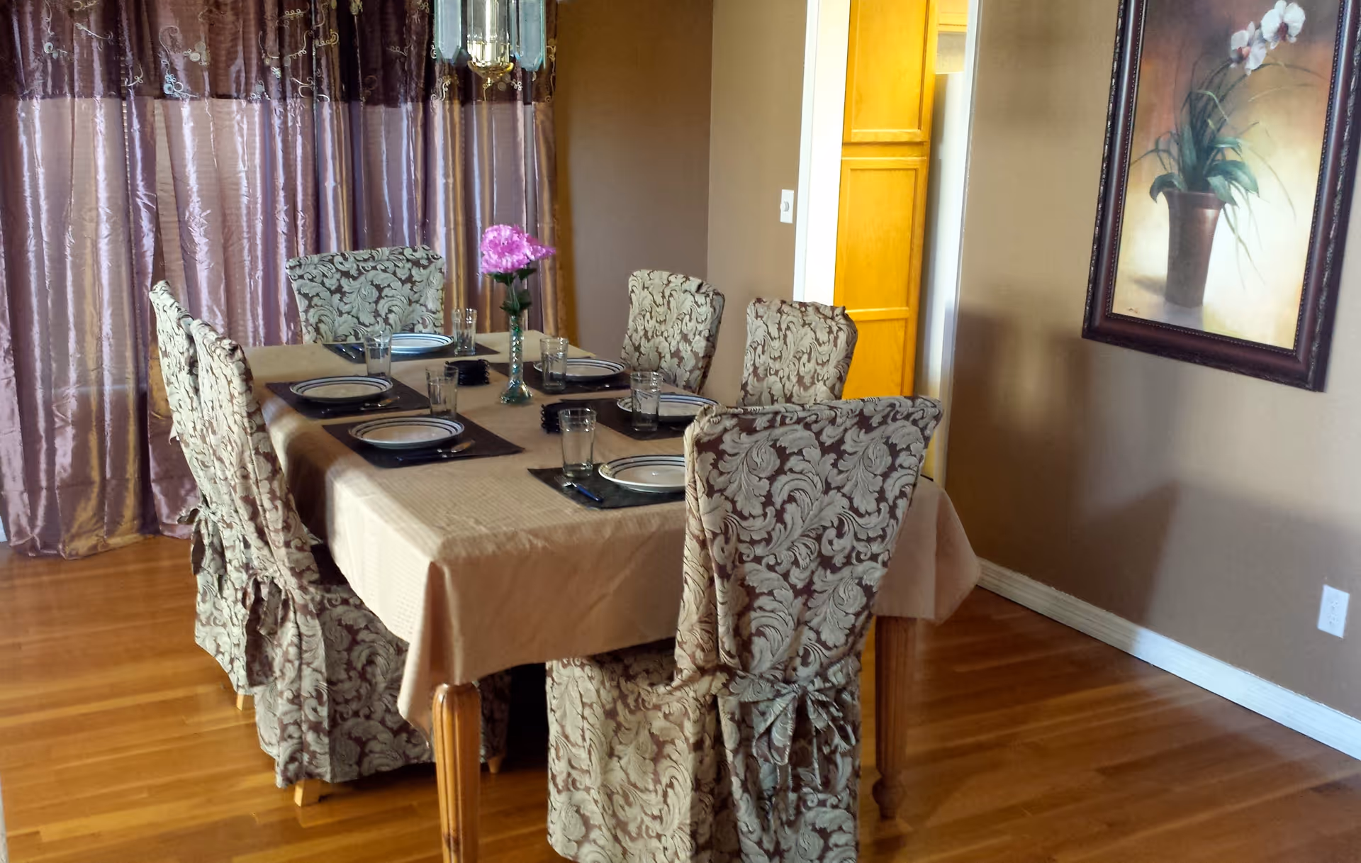 A dining room with a table set for six, patterned slipcovered chairs, a pink flower centerpiece, curtains and framed wall art.
