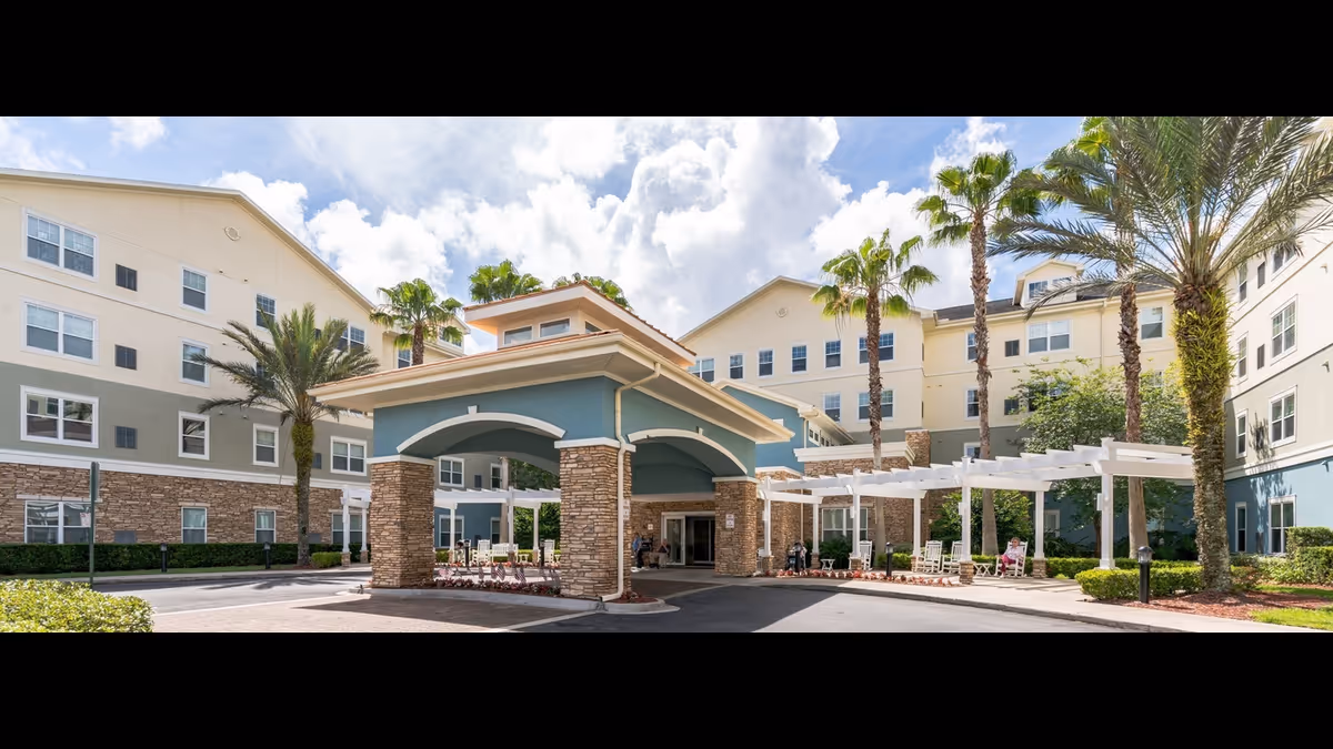 Front entrance of a multi-story senior living facility with a covered porte-cochere, palm trees, and outdoor seating.