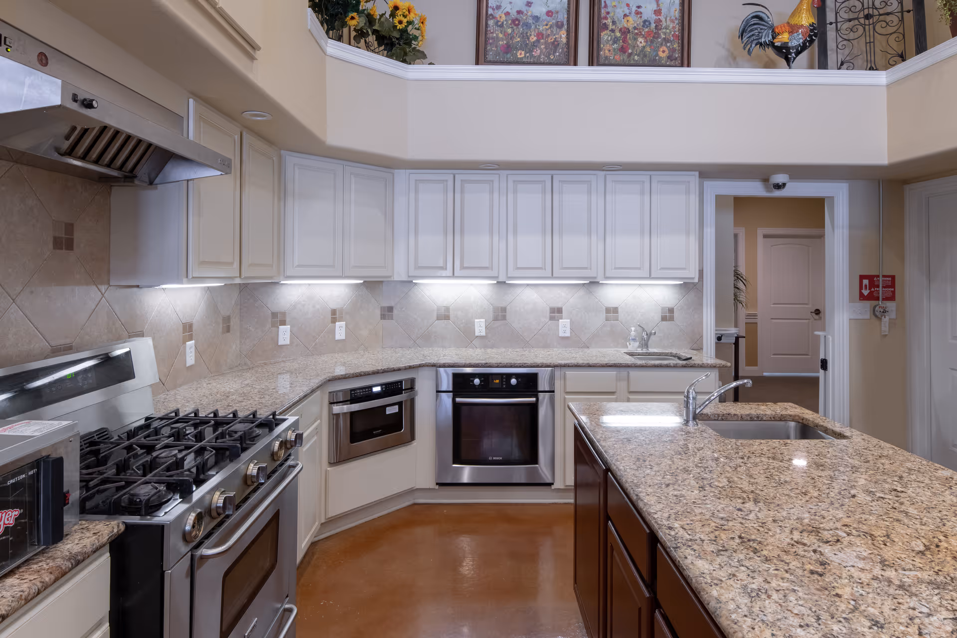 A modern kitchen with granite countertops, white upper cabinets, and stainless steel appliances including a stove, oven, microwave, and sink. The kitchen has a large island with a sink and faucet, and the walls are decorated with beige tiles. Above the kitchen area, there is a second-floor railing with floral artwork and a decorative rooster statue.