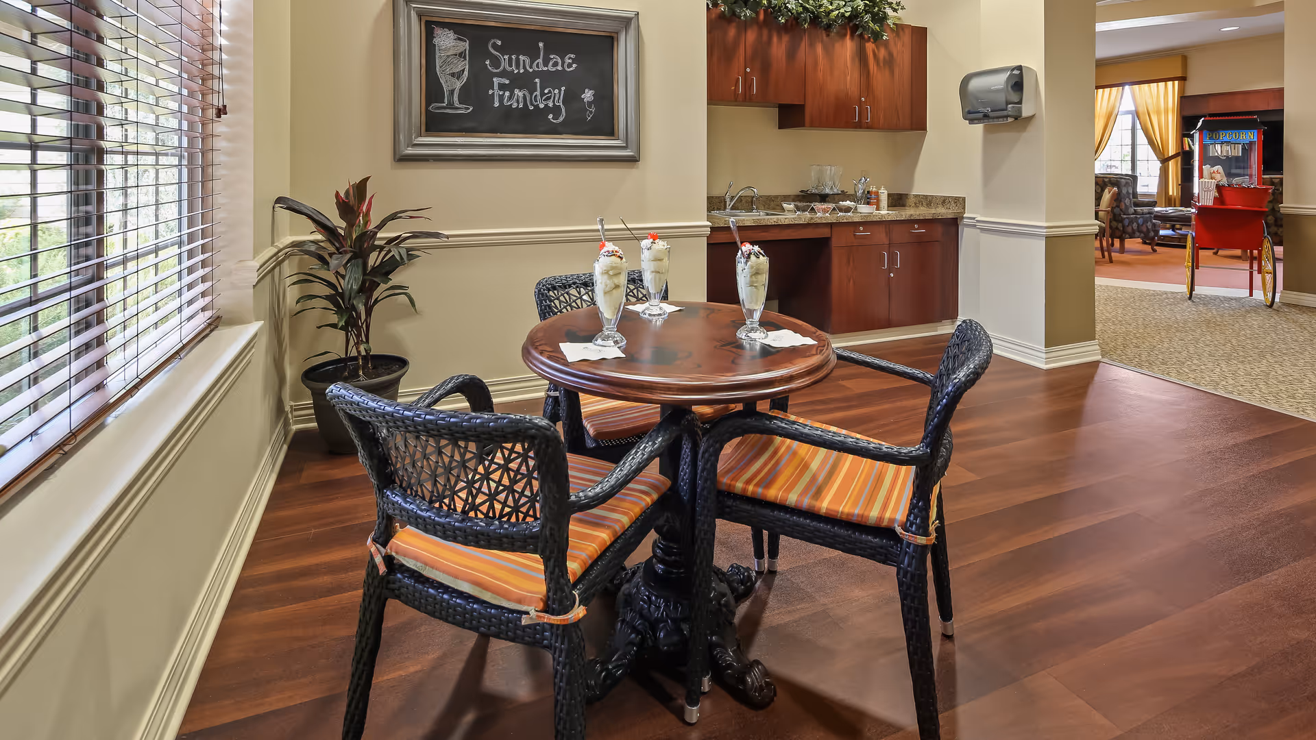 A cozy indoor seating area with a round wooden table and four wicker chairs with orange striped cushions. On the table are three ice cream sundaes with spoons. Behind the table is a small kitchenette with a granite countertop, wooden cabinets, and a paper towel dispenser. A chalkboard on the wall reads 'Sundae Funday'. To the right, there is an open area with a popcorn machine and additional seating visible in the background. Large windows with blinds allow natural light into the space.