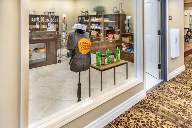 View through a glass window into a small retail pantry area in a senior living facility. Inside, shelves are stocked with various packaged goods and snacks. A mannequin displays a gray t-shirt with an orange circle and the text 'LEGACY LOVES ORANGE'. A small table holds several green water bottles and travel mugs. The hallway outside has patterned carpet and beige walls.