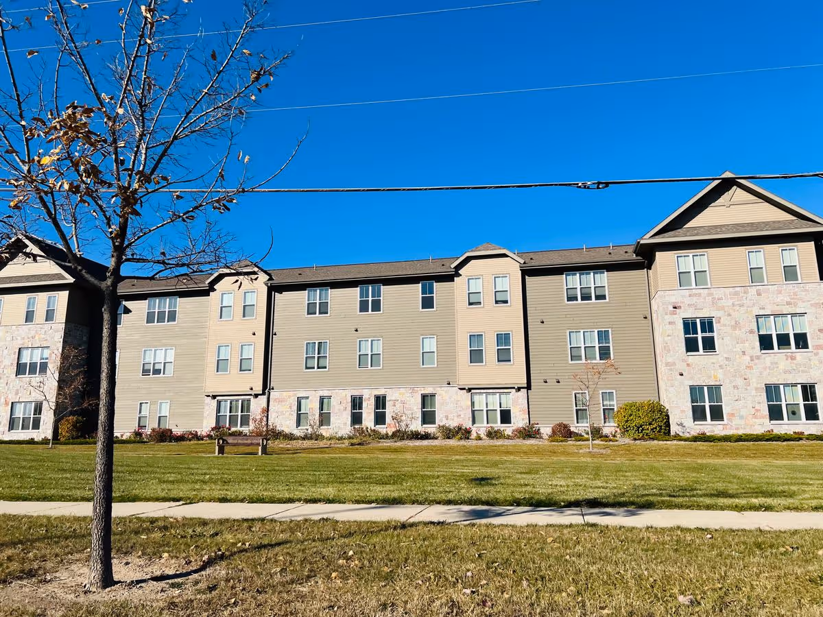 Front exterior of a three-story residential building with a grassy lawn, sidewalk, a bench, and a bare tree under a clear blue sky.