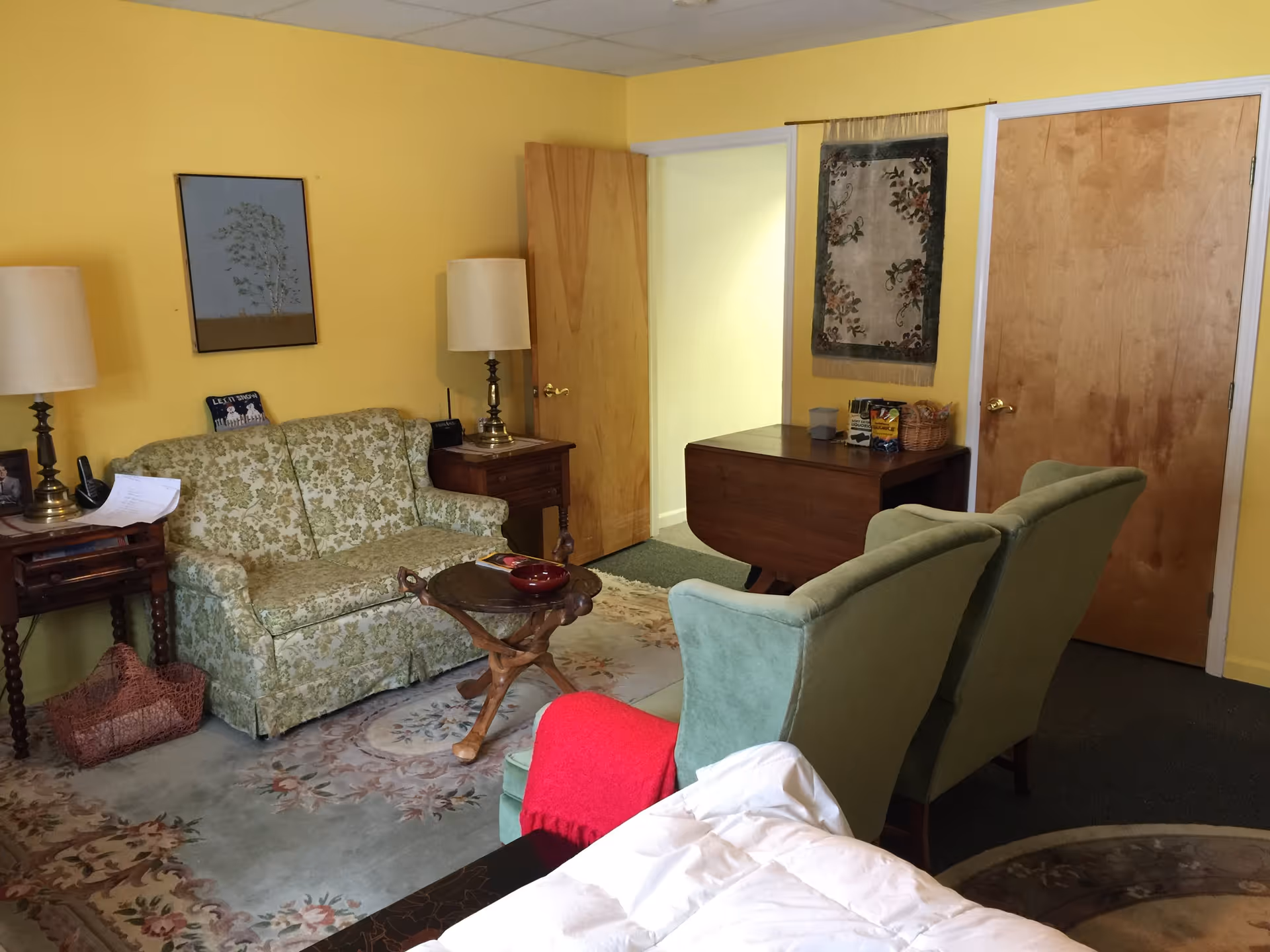 Cozy sitting area with a floral loveseat, two green armchairs, small tables and lamps against yellow walls.